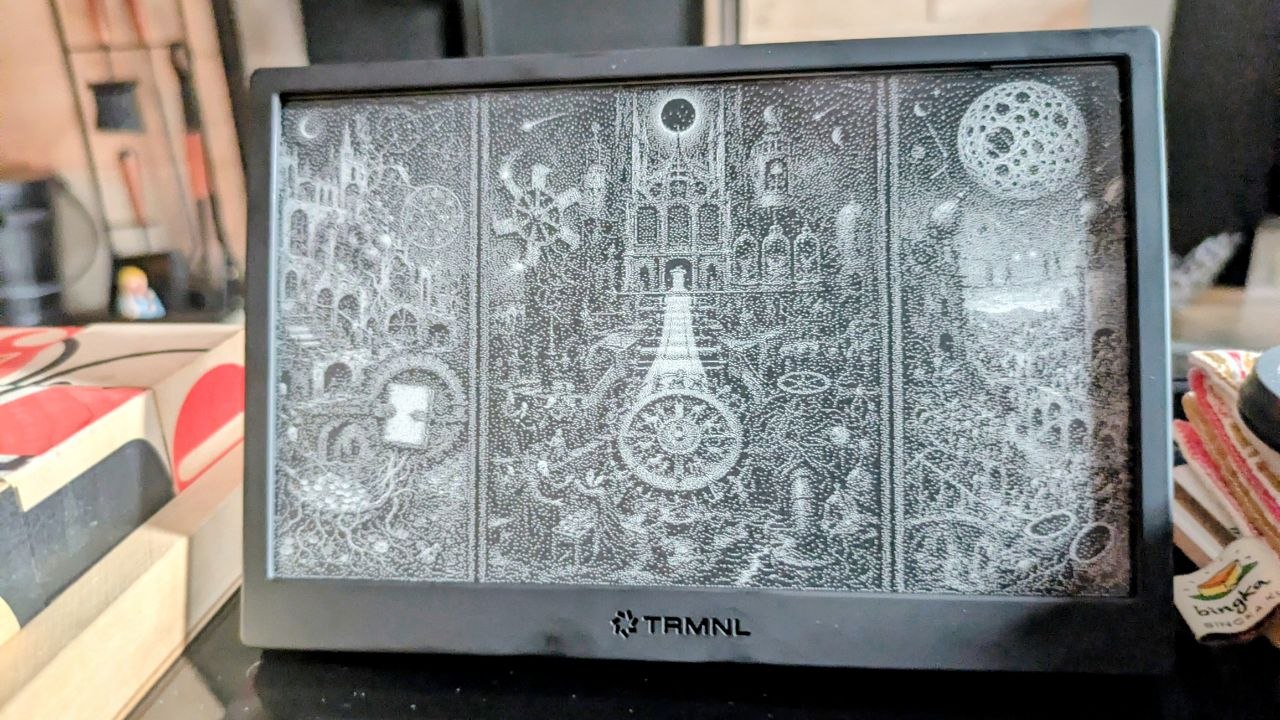 A TRMNL e-ink display on a desk showing a black-and-white Daily Bosch triptych, with books and small objects nearby.